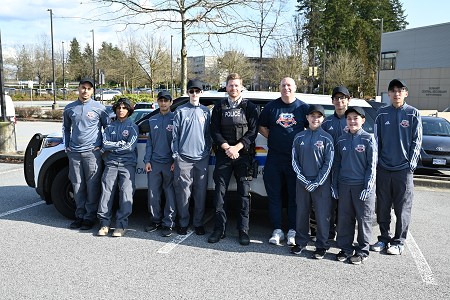 A group of 7 Youth Academy participants standing in front of a police vehicle with two Burnaby RCMP police officers. One police officer is in uniform and the other is a Youth Academy instructor.