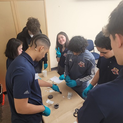 A group of 7 Youth Academy participants standing around a table, wearing blue gloves, while working with fingerprinting powder.
