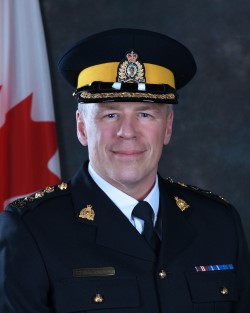 Le surintendant principal Graham de la Gorgendiere, vêtu d’un uniforme bleu, sourit devant un drapeau du Canada
