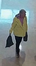Une femme aux cheveux gris courts portant une veste jaune vif et un pantalon noir. Elle porte deux sacs noirs, un dans chaque main.