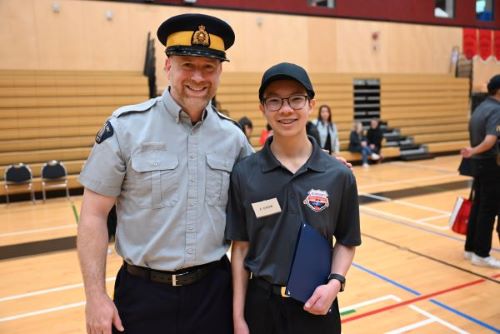 A police officer in an RCMP uniform stands beside a smiling teenager inside a school gymnasium