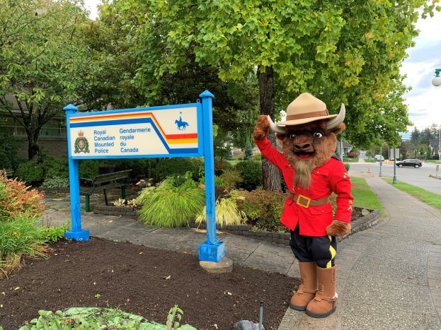 Une mascotte de bison vêtue d’une tunique rouge officielle de la GRC, portant des bottes brunes et un chapeau Stetson brun, faisant un salut d’une main et se tenant à côté d’un panneau de la GRC.