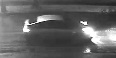 A night time, black and white photo of a vehicle on the roadway with its headlights on.