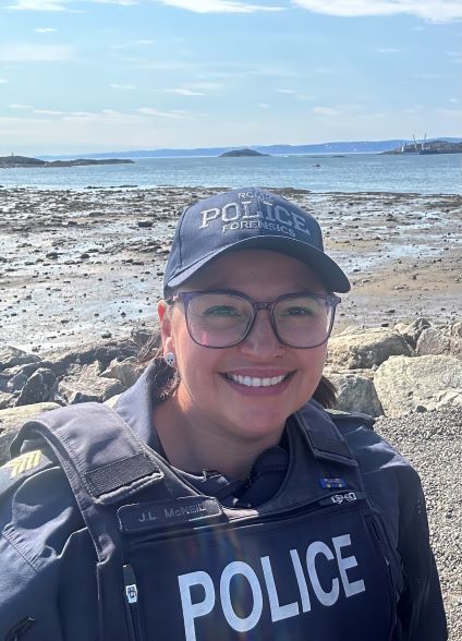 RCMP police officer is wearing a blue police uniform and blue baseball cap with RCMP police written on it while standing on a  rocky shoreline in Nunavut.