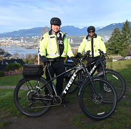 Deux policiers portant une veste réfléchissante se tiennent avec des vélos électriques devant eux et des montagnes en arrière-plan. 