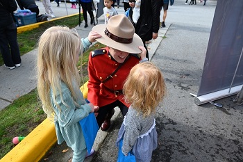Une gendarme en tunique rouge s’agenouille pendant que deux petites filles jouent avec son chapeau Stetson 