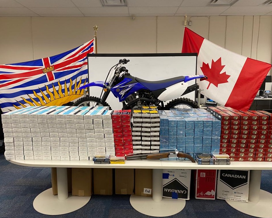 Table stacked with dozens of cartons of contraband cigarettes, a sawed off shotgun and ammunition with a motorcycle behind it. All seized by Burnaby RCMP's Prolific Offender Suppression Team.
