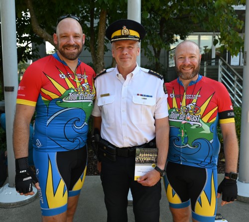 The Wheeler brothers, dressed in cycling attire stand on either side of Burnaby RCMP's Officer In Charge on a sunny day outside Burnaby City Hall. 