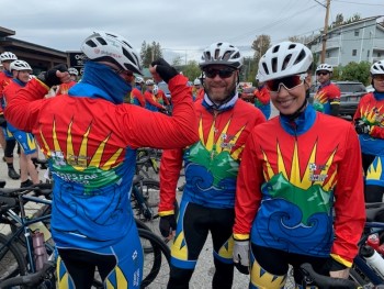 A man in cycling attire with his back to the camera beside Staff Sgt. Rob Wheeler and Sgt. (Ret) Tess Landry, who are smiling in cycling jerseys, helmets, and sunglasses. 