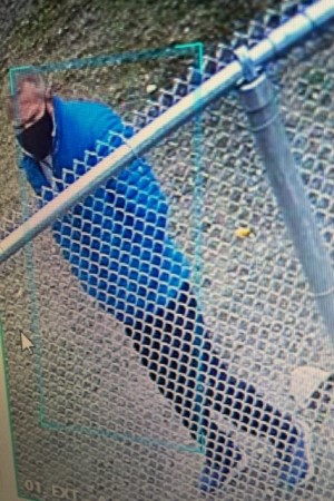 Left side view of a different view of a person of interest stands near a fence during the daytime. He is wearing a black medical mask and blue jacket.