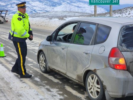 Un policier portant une veste de sécurité jaune s’approche d’un véhicule gris immobilisé sur une autoroute enneigée. 