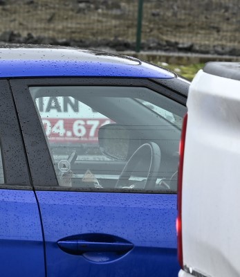 A hand on a cell phone close to a steering wheel inside a blue vehicle 