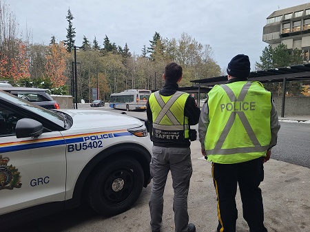 The backs of two men. One wearing a police vest, the other wearing an SFU Campus Public Safety vest. they are standing beside a Burnaby RCMP Police car at SFU