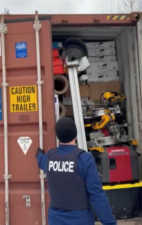 A police officer stands outdoors near the open door of a red shipping container. The container is packed with various items and boxes. 