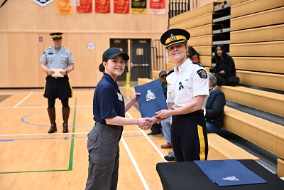 Inspector Peri Mainwaring presenting a youth academy participant with a certificate in a blue folder, with RCMP logo, both are smiling.