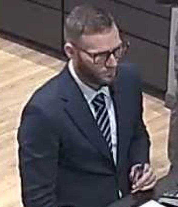 Caucasian man with short light brown hair and short beard, wearing a dark suit jacket with a striped tie, glasses and white dress shirt.