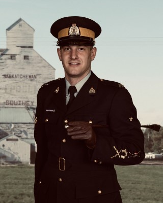 Cpl. Kyle Kifferling, in a blue uniform and hat, smiles in front of a large barn emblazoned with the name of his hometown, Southey, Saskatchewan