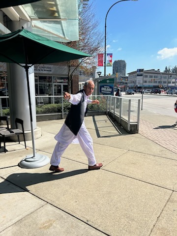 South Asian man, balding with grey hair wearing a grey vest overtop of a long sleeve white linen shirt, matching white linen long pants and brown dress shoes, walking out of a restaurant with his arms out.