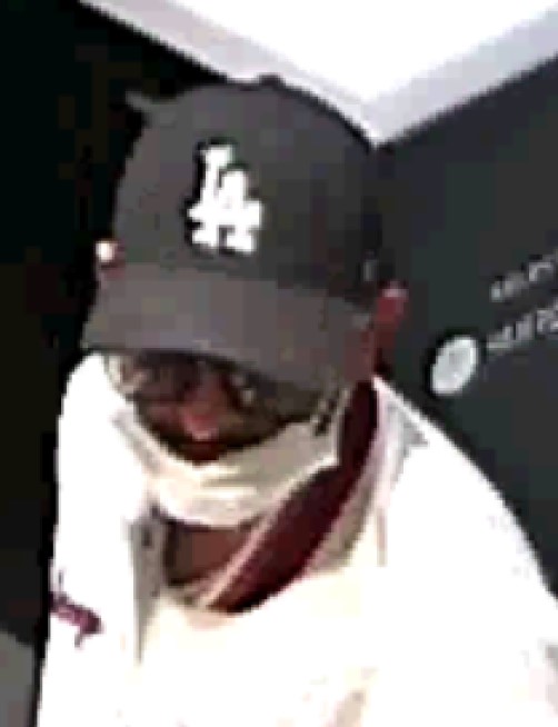 A man wearing glasses, a white medical mask, dark LA Dodgers baseball hat and a white bomber jacket.