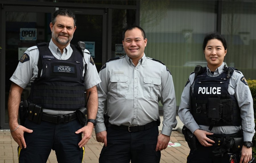 Two male RCMP officer stand  smiling beside Cst. Shaelyn Yang outside an office building