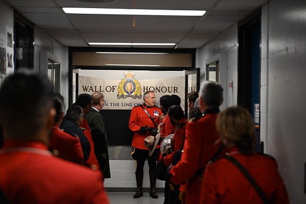 Burnaby RCMP Officer in Charge, Chief Superintendent Graham de la Gorgendiere, stands in a Red Serge beside the Burnaby RCMP Wall of Honour alongside a group of people 