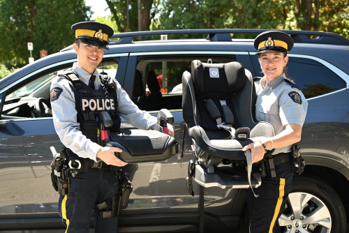 Two police officers in uniform stand near a vehicle outdoors while holding a car seat and a booster seat