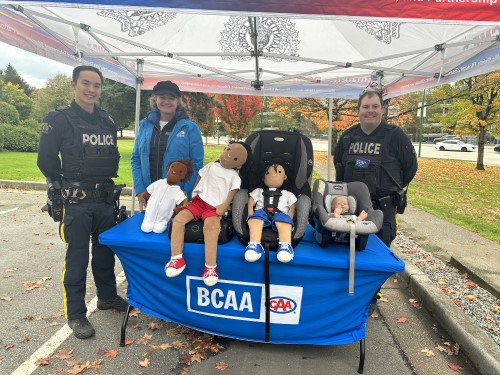 Two police officers in uniform stand outdoors with a BCAA employee. On a table in front of them are several car seats and child-sized dolls. 