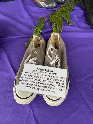Pair of women’s shoes placed on purple cloth with a biography placard that says, </q>Helene Colgan. Died: December 6, 1989. Helene (23) was in her final year of the mechanical engineering program and planned on getting her master’s degree. She had three job offers waiting. Helene was one of 14 women killed at Ecole Polytechnique in Montreal.</q> 
