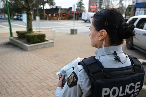 A female police officer in uniform stands on a sidewalk beside a street holding a plastic bag filled with cold weather items such as gloves and socks